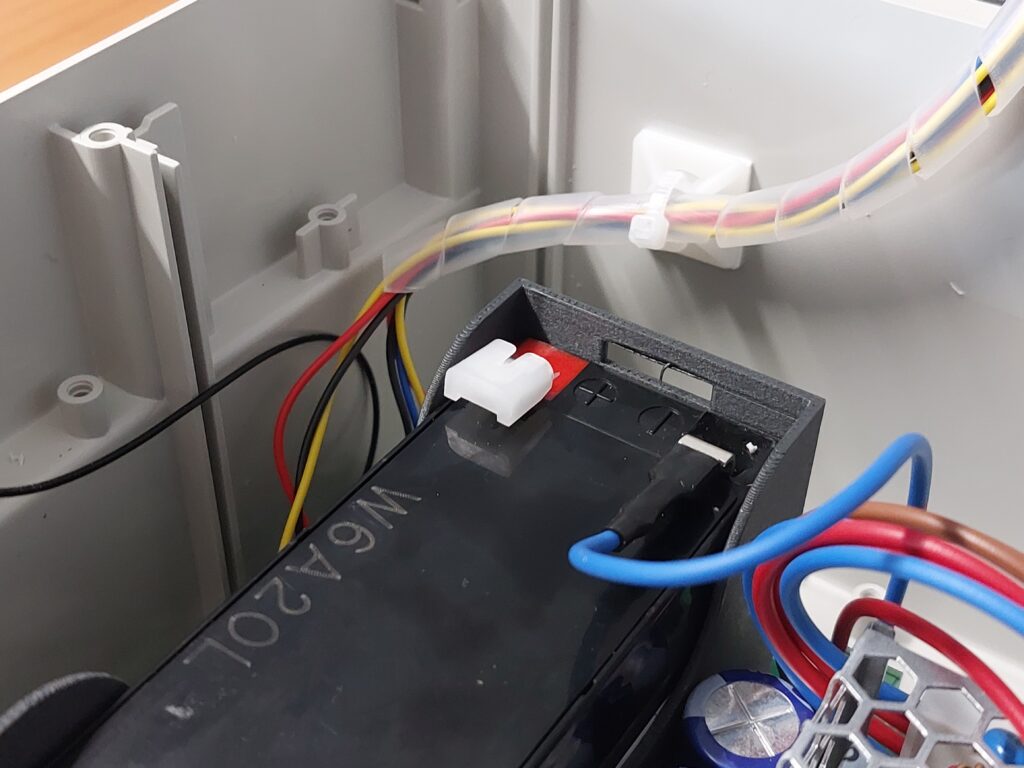 A close-up of the black, rectangular battery (W6A20L) mounted on the motherboard. The battery’s metal contacts still have their protective red-and-white plastic caps on. The power cables lie loosely, ready to be connected.