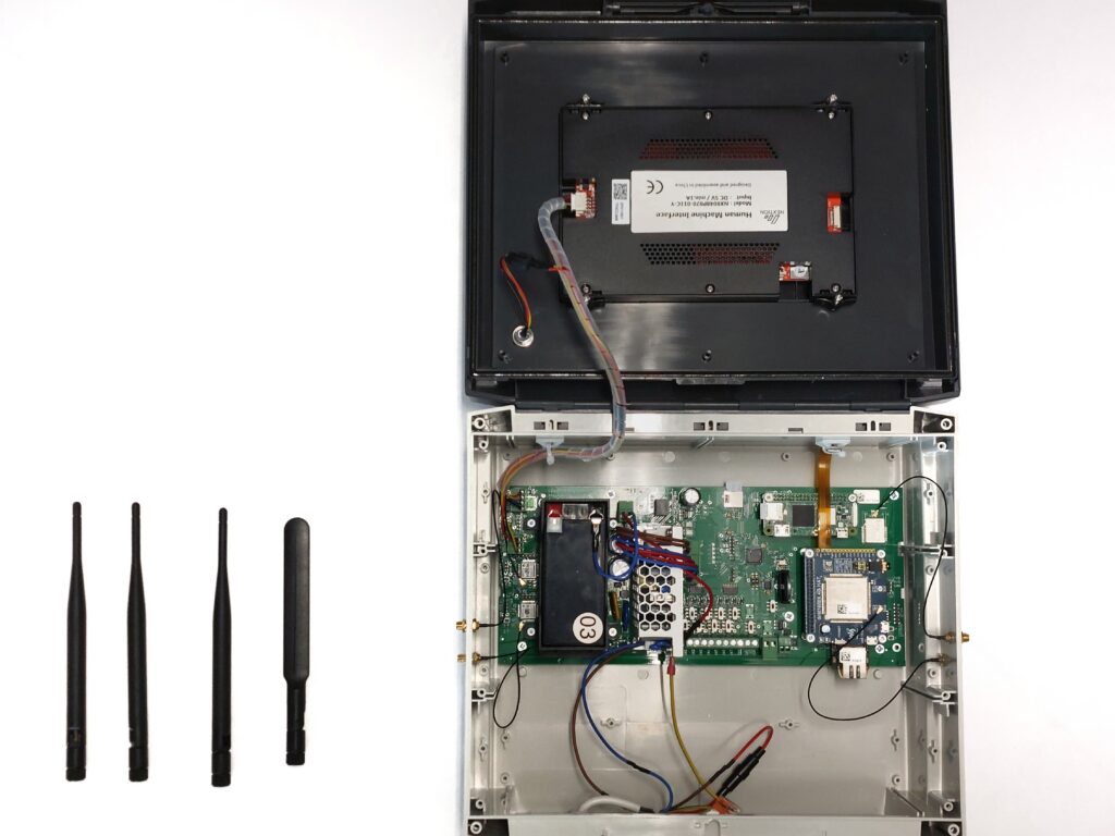A photo of the device with the case open (top view of the interior). Next to the case lie four neatly arranged black antennas ready for installation—three of them are slender and identical, while one is noticeably shorter and flatter (intended for the SIM module). Inside the device, you can see, among other things, the battery and the green motherboard.