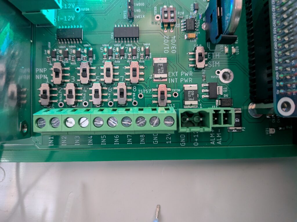 Zbliżenie na dolną część zielonej płyty głównej (PCB) urządzenia FarmWatch Pro. Na dole znajduje się długa, zielona listwa zaciskowa ze złączami opisanymi jako wejścia cyfrowe od IN1 do IN8, a także stykami masy (GND) i zasilania (12V). Powyżej listwy umieszczono sekcje fizycznych przełączników suwakowych służących do sprzętowej konfiguracji parametrów pracy tych wejść.