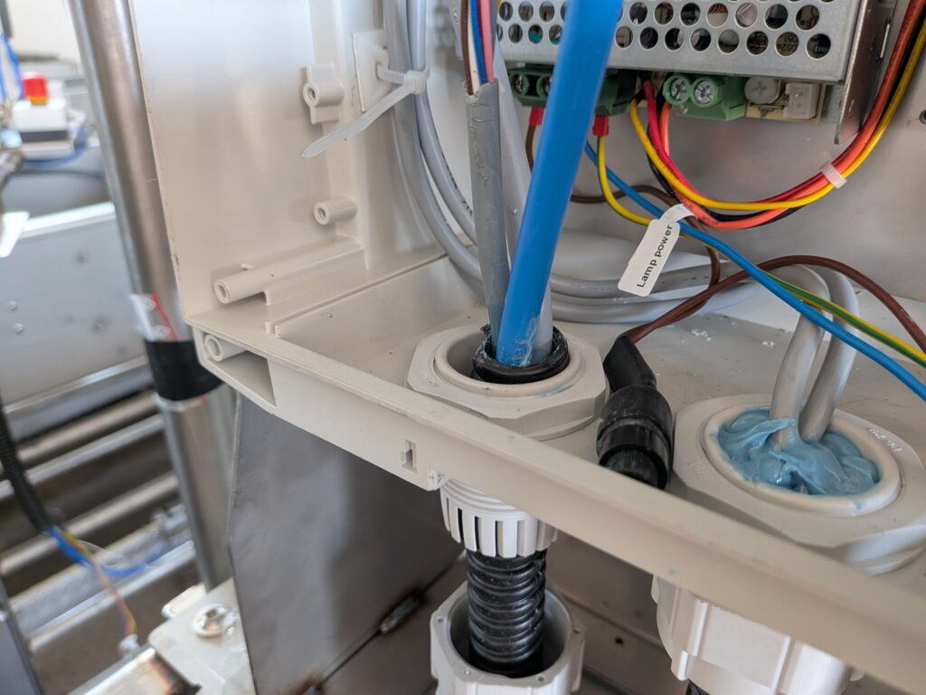Wnętrze obudowy sterownika z górnym przepustem kablowym, przez który przeprowadzono niebieski przewód pneumatyczny oraz przewody zasilania oznaczone opisem Lamp power.